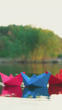 several multi-colored origami in the shape of ships are floating on the water in the lake in the evening on the background of the summer landscape. vertical video