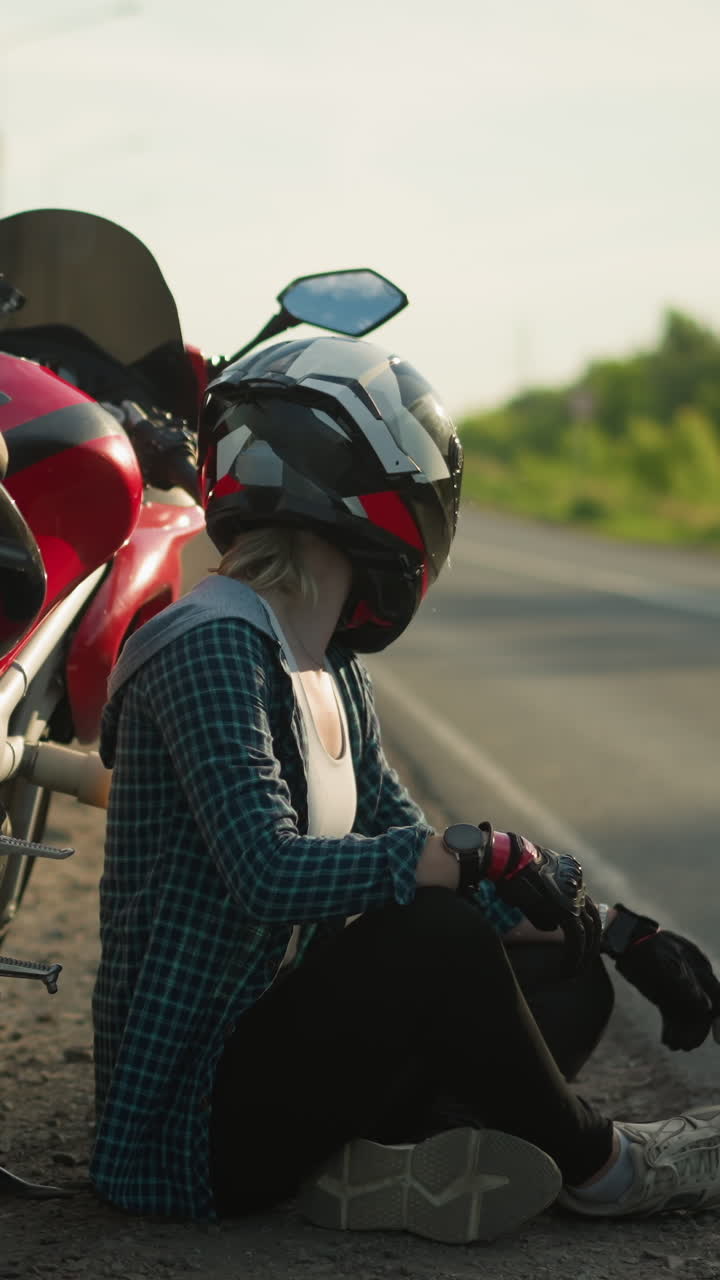 헬을 입은 여성 오토바이 운전자는 시골 도로 에 주차된 빨간색 파워바이크 에 바닥에 다리를 어 앉아 평온하게 주변을 관찰하는 것처럼 보입니다.