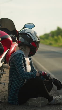 una motociclista femenina, con casco, se sienta con las piernas cruzadas en el suelo al lado de su bicicleta eléctrica roja estacionada junto a una carretera rural, parece estar observando con calma su entorno