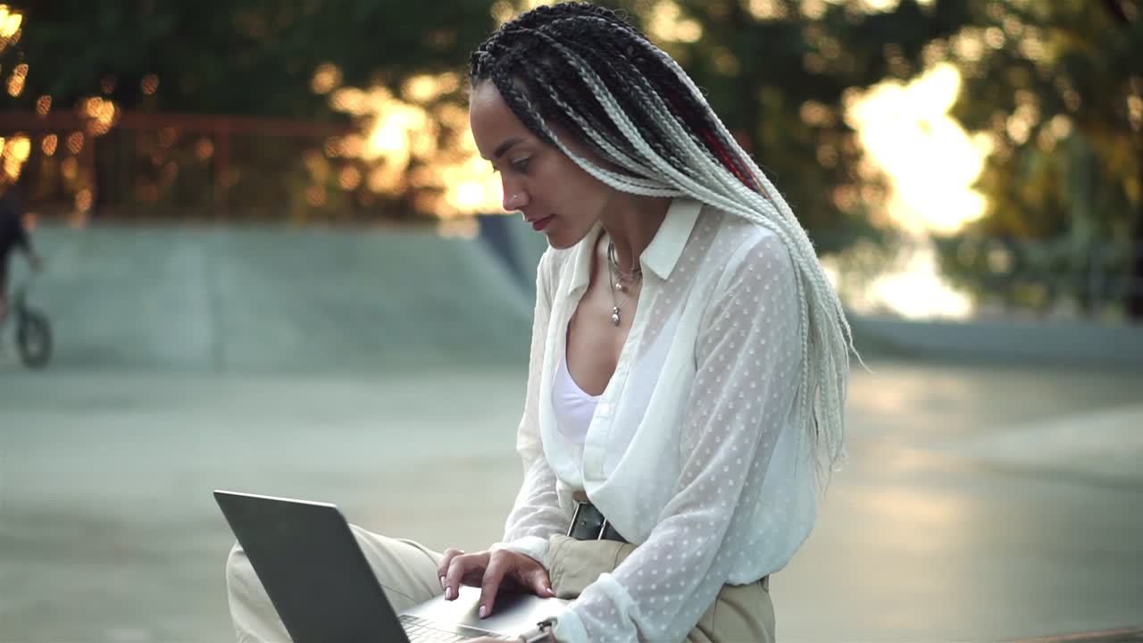 Side view of beautiful, elegant girl with black and white dreadlocks sitting on parapet in local skatepark working on laptop