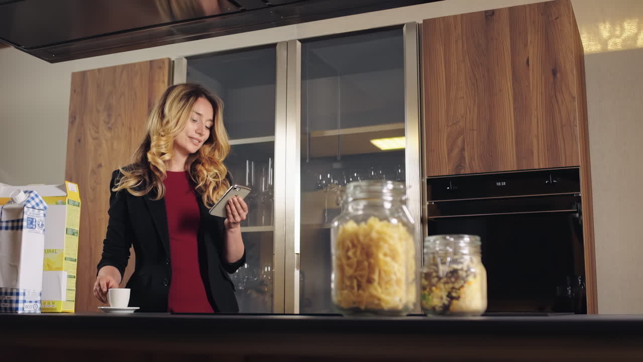 CHISINAU, MOLDOVA - SEPTEMBER 25, 2018: A smiling caucasian woman with blonde hair in jacket drinking coffee and looking at the phone on a kitchen. Table with food