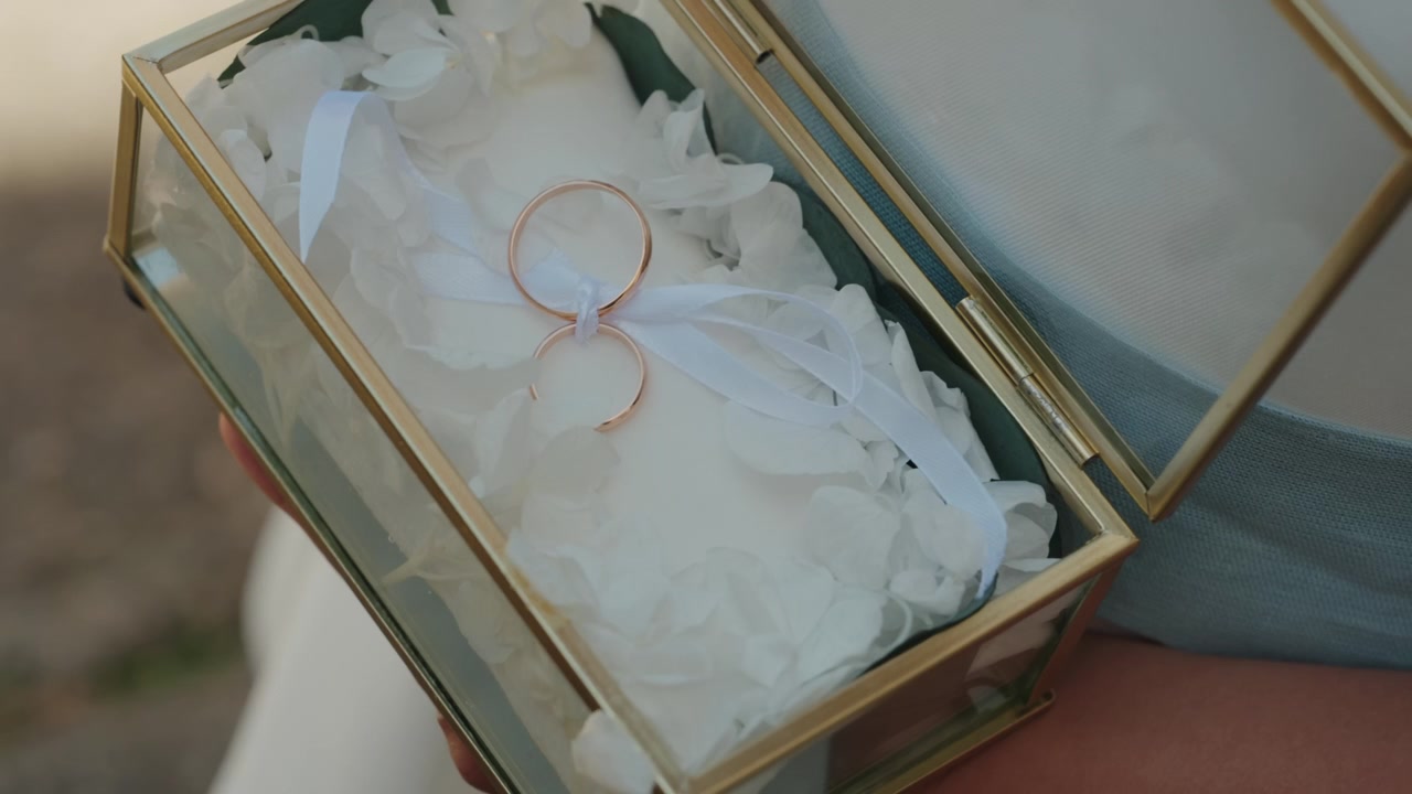Two gold wedding rings bound with ribbon resting on white petals in glass and brass box