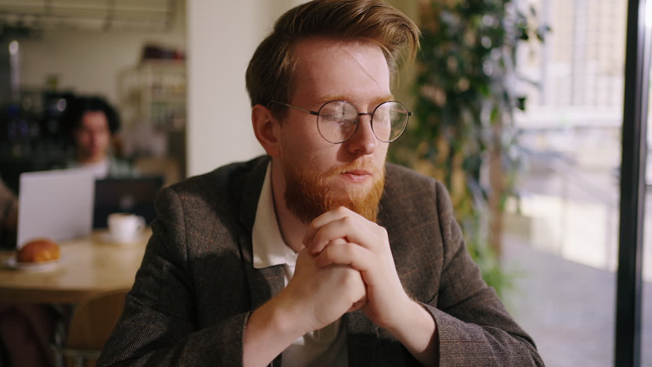 A young man with a beard and glasses sits in a coffee shop, looking away and thinking