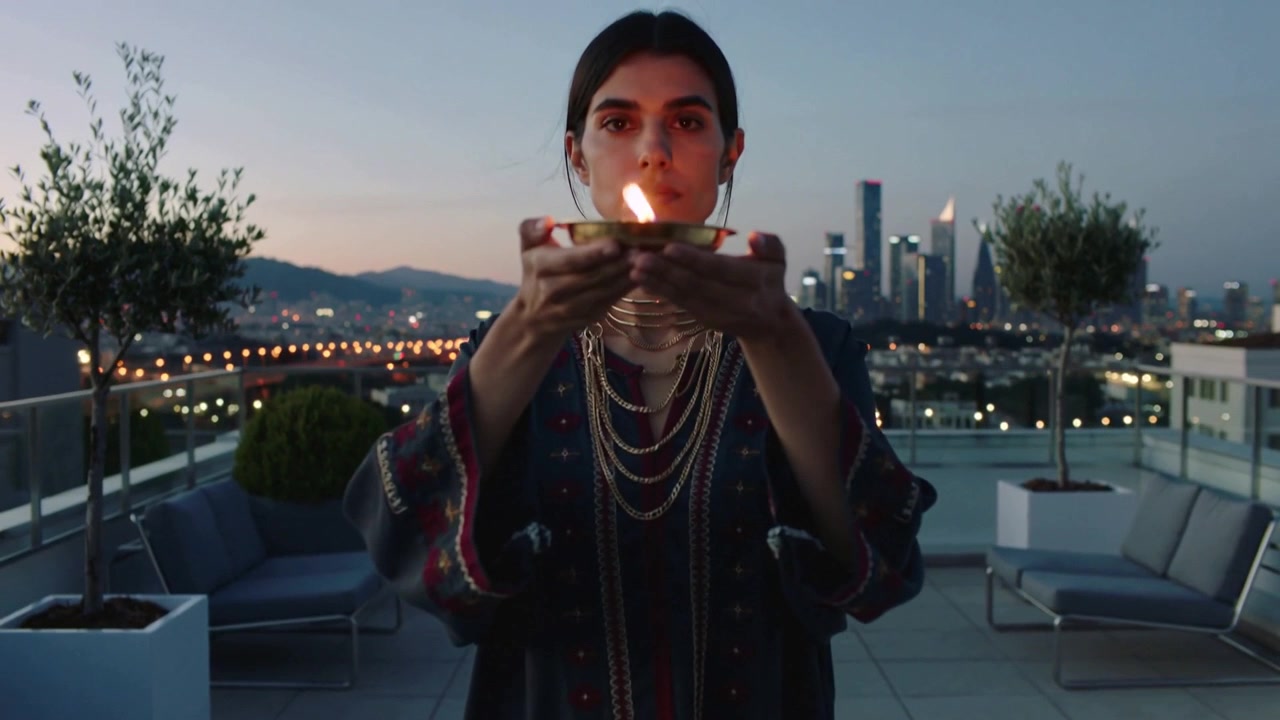 Dusk falling, woman wearing dark robe and gold-chokers raising brass lamp on rooftop offering light