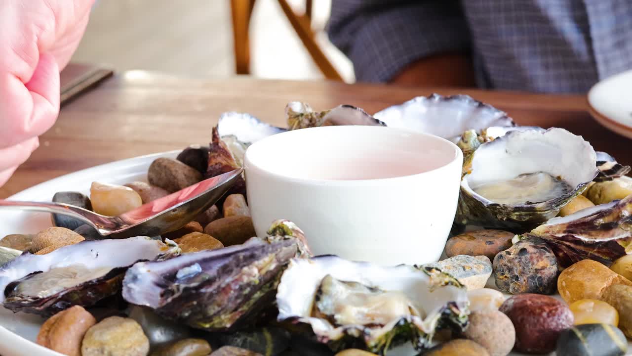 Hands prepare oysters with sauce on a stone-decorated plate in natural light, creating a casual dining atmosphere