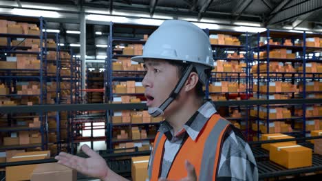 close up side view of asian male engineer with safety helmet standing in the warehouse with shelves full of delivery goods. wondering and saying why working doubtfully in the storage