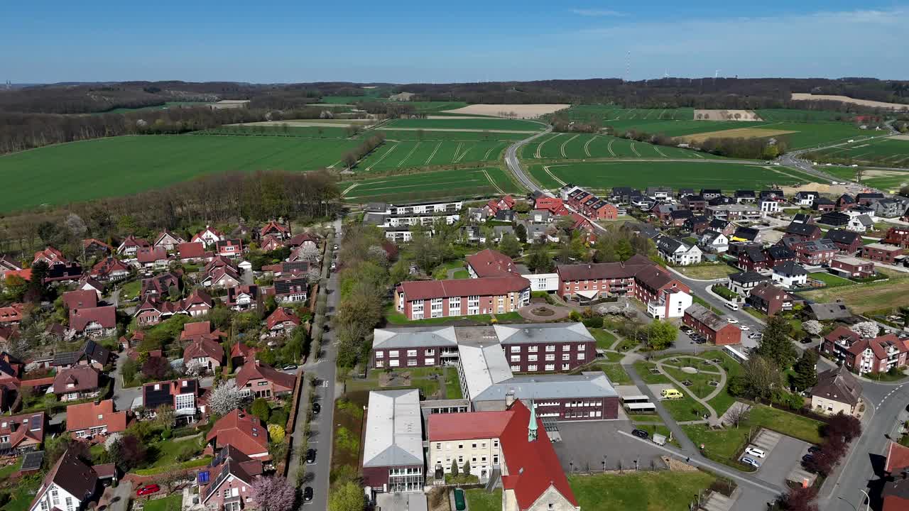 Aerial flyover small german city with hospital building and rural countryside fields. Wide shot. Green farm fields and hills in Background. Sunny quiet and calm spring day in America, USA.