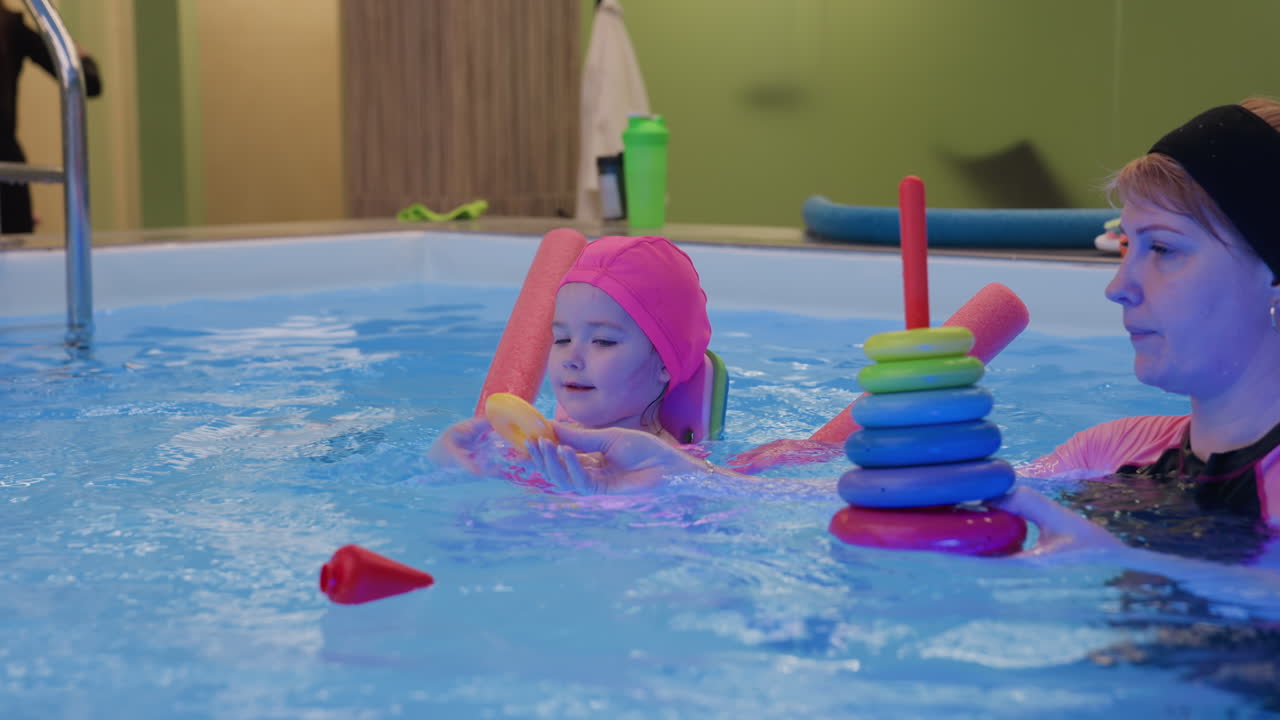 Swim coach holding pyramid toys then throwing into water for fun, bright pool light, playful training cue, gentle splash, safe supervised moment, blue ripples, child ready to chase shapes