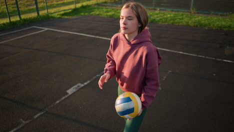 young athlete with maroon hoodie bouncing volleyball, preparing to serve in an outdoor court with greenery in the background and clear court markings visible on the ground