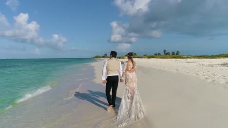 Pareja-Adulta-Y-Novia-Caminando-Tomados-De-La-Mano-En-La-Playa-De-Arena-Blanca,-Hombre-Señalando-El-Mar-Desde-Atrás