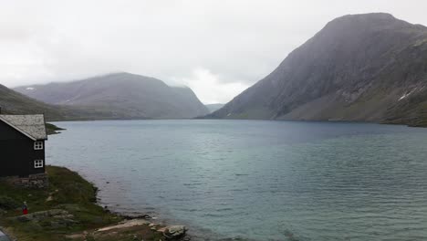 Gran-Lago-Azul-En-Medio-De-Una-Llanura-En-Una-Cadena-Montañosa-Rocosa-Con-Una-Casa-Grande-Y-Algunos-Turistas,-Noruega,-Europa,-Drone