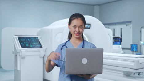 asian female doctor smiling and pointing to a laptop in hand while standing with mri machine