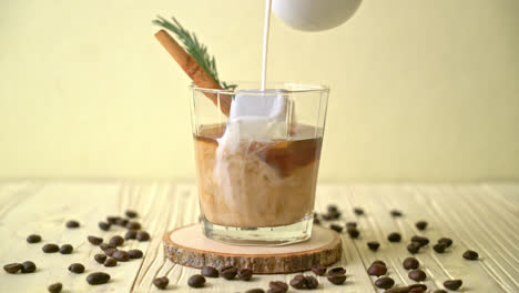 pouring-milk-in-black-coffee-glass-with-ice-cube,-cinnamon-and-rosemary-on-wood-background