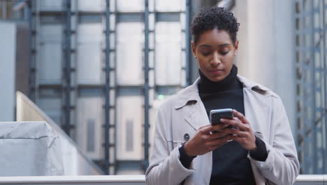 Close-up-of-fashionable-young-black-woman-standing-in-the-city-in-front-of-a-modern-building-using-her-smartphone,-front-view,-waist-up