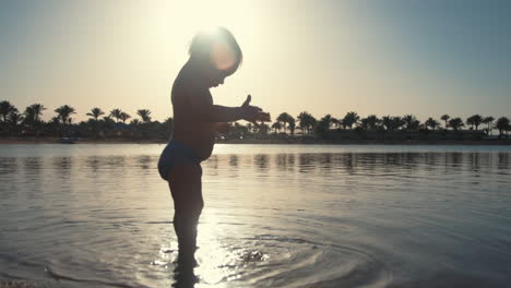 Lächelnder-Kleiner-Junge-Wirft-Sand-Ins-Meerwasser-An-Der-Küste-Bei-Sonnenuntergang.