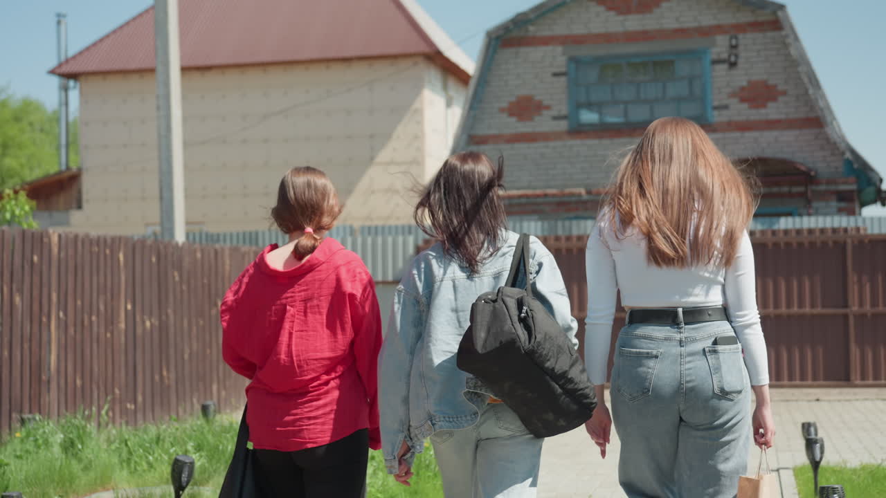 Three friends walk out compound through open gate, one in red jacket holds arm around friend neck while carrying tote bag, passing paved area under clear blue sky near wooden fence and green grass
