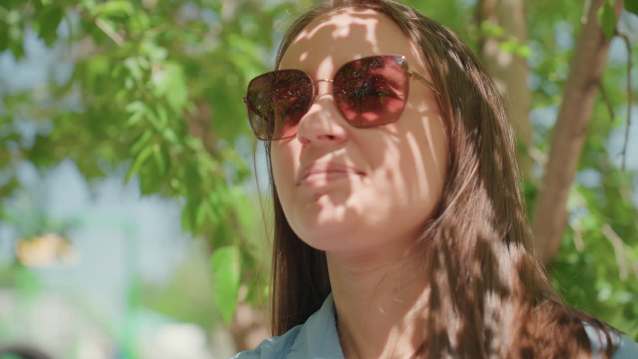 Освещенная солнцем женщина в солнечных очках под деревом улыбается, а пятнистый свет играет на ее лице, джинсовая рубашка, лиственный парк на заднем плане, теплое летнее настроение, видеоролик в стиле непринужденного образа жизни, демонстрирующий расслабленный портрет на открытом воздухе