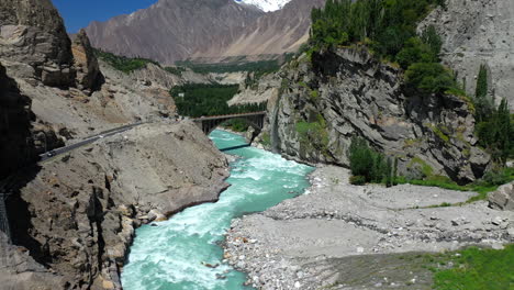 drone shot of turquoise blue water river flowing under a bridge in karakoram mountain range along karakoram highway, moving through mountain pass towards bridge