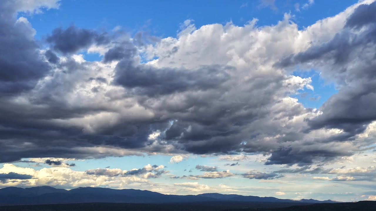 4K drone timelapse of thick, dark clouds rolling in to cover the blue sky. The fading sunset light casts a warm glow near the horizon above a silhouetted mountain range as evening approaches.