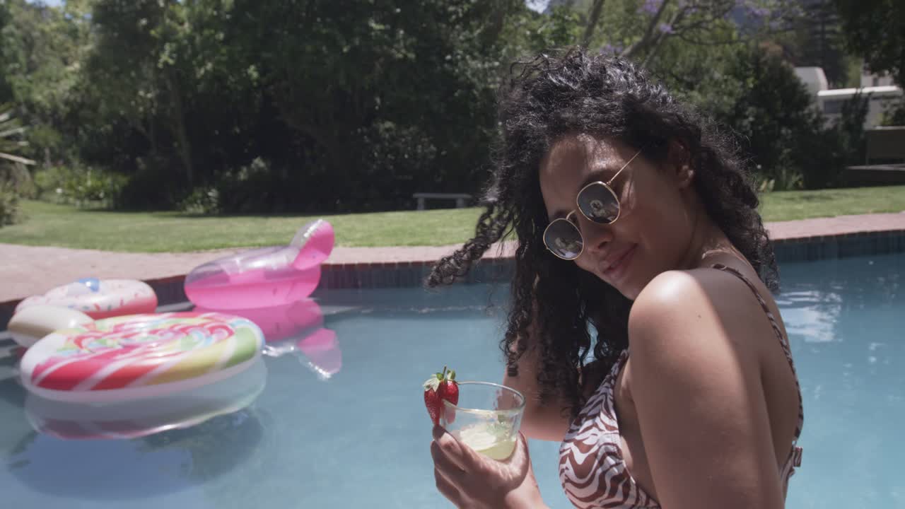 African American woman seeing camera poolside flipping hair sipping strawberry cocktail, copy space