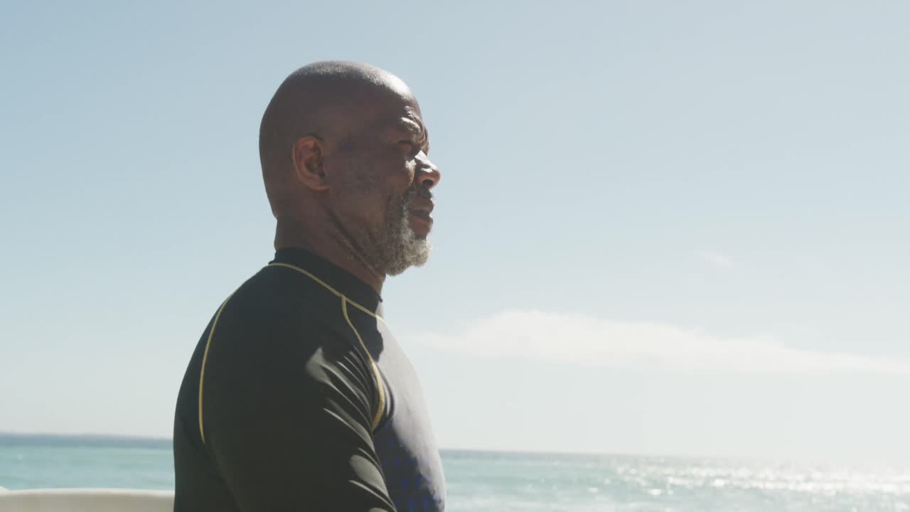 Portrait of smiling senior african american man walking with surfboard on sunny beach