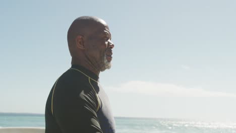 Portrait-of-smiling-senior-african-american-man-walking-with-surfboard-on-sunny-beach