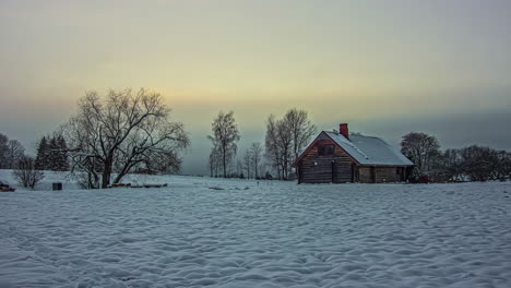 Static-shot-of-beautiful-wooden-cottage-covered-with-thick-layer-of-white-snow-with-sun-setting-in-timelapse-in-the-background-at-dusk
