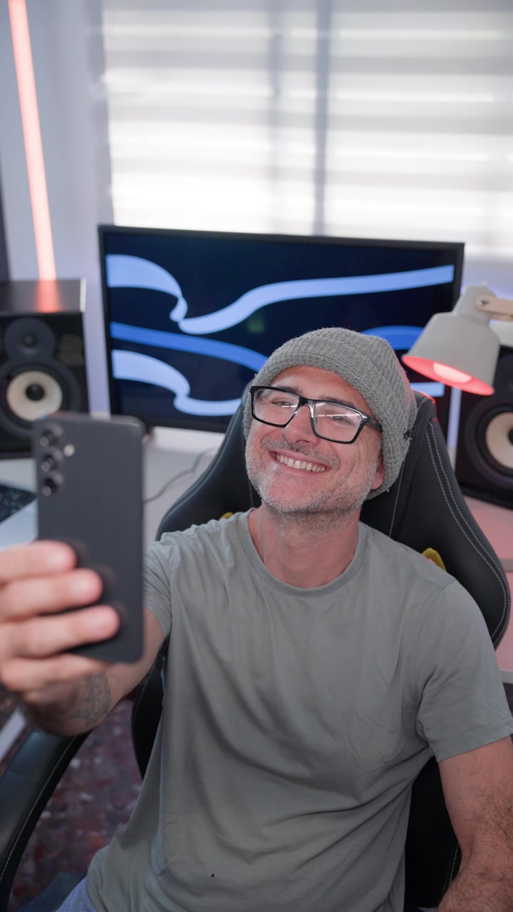 Un creatore di contenuti che indossa un berretto e occhiali sorride mentre si scatta un selfie verticale con il suo telefono in uno studio casalingo, Verticale