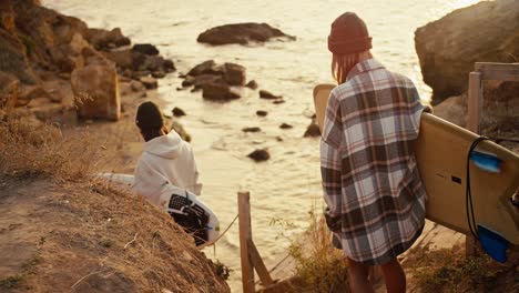 un hombre moreno en una sudadera blanca y una mujer rubia en una camisa a cuadros están llevando tablas de surf mientras bajan una escalera de madera a una orilla rocosa cerca del mar al amanecer en verano en verano