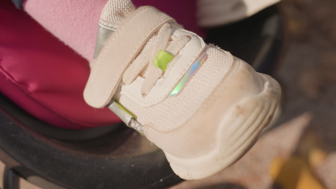 Primo piano di una scarpa da neonato sul passeggino, scarpa da ginnastica in velcro sul poggiapiedi con calze rosa pastello, suola consumata e striscia riflettente, foglie del parco sotto i piedi, movimento delicato, atmosfera accogliente di gita in famiglia.
