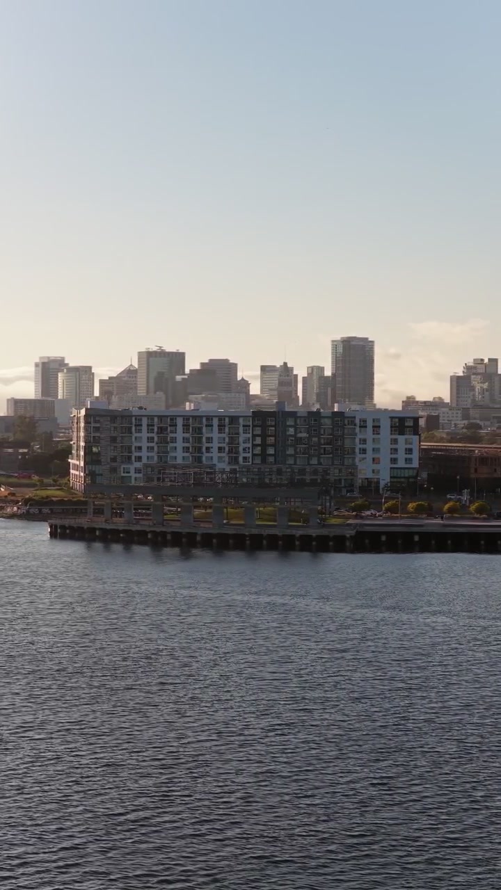 Imagens aéreas de drone voando sobre a água em direção aos edifícios modernos e ao horizonte da cidade de Oakland, Califórnia, com a baía calma e um céu claro durante um belo e quente pôr do sol