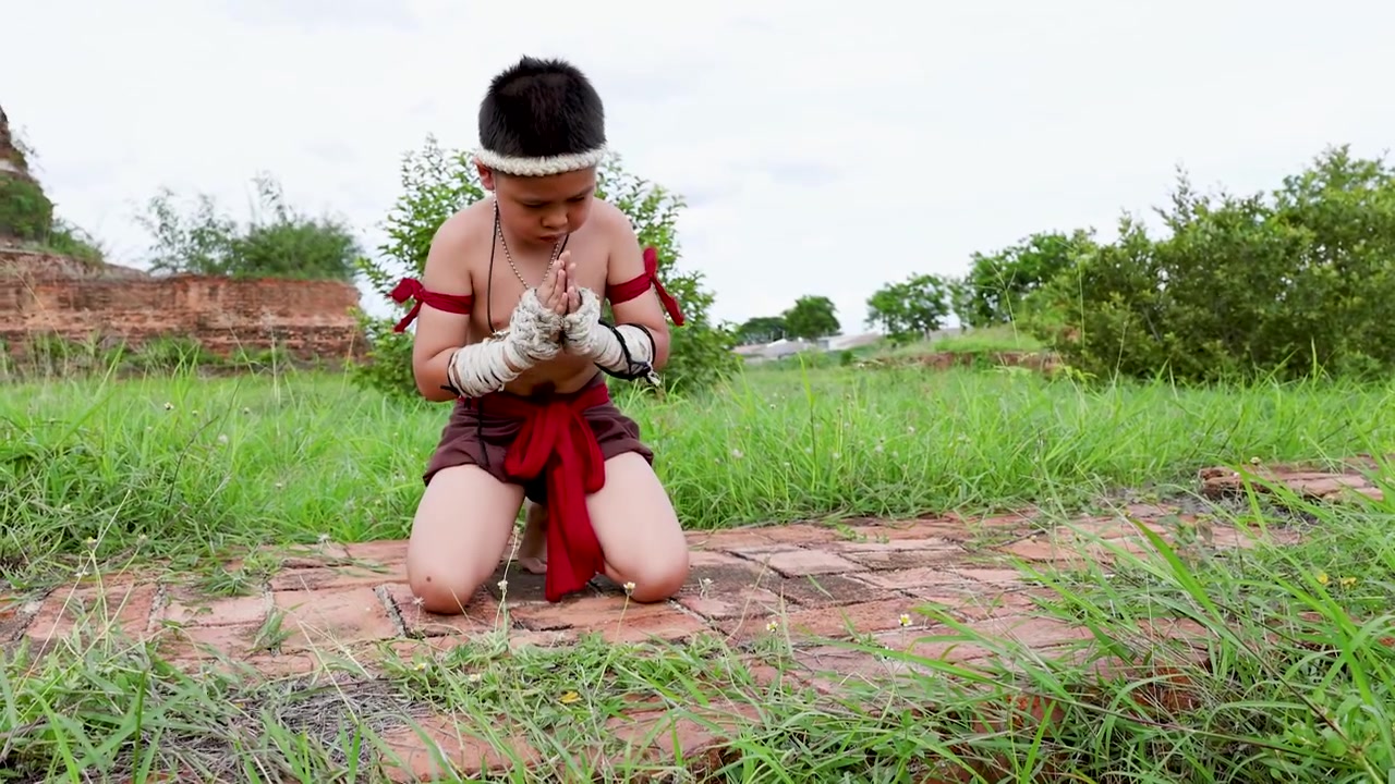 Bambino asiatico esegue una preghiera rituale in un costume tradizionale di arti marziali vicino alle rovine di un'antica pagoda di pietra