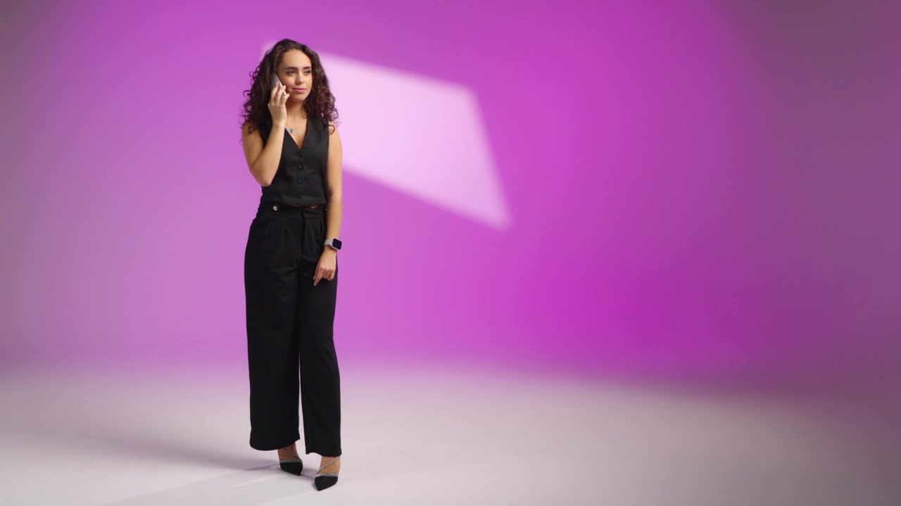 Scatto a figura intera di un'elegante donna sportiva con gilet nero, pantaloni a gamba larga e tacchi che parla al telefono, sorride e gesticola su uno sfondo studio rosa-viola
