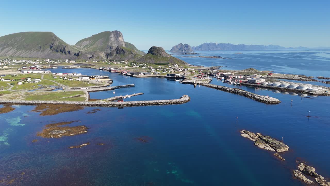 Aerial view of Værøy harbor: colorful village by the sea, dramatic mountains rising behind, and the endless blue of the Norwegian coast
