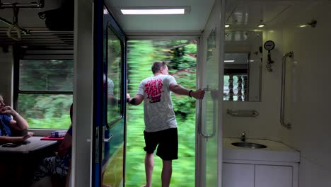 an adventurous tourist enjoys a thrilling view by hanging out of a moving train door in sri lanka, experiencing the lush greenery during a memorable journey.