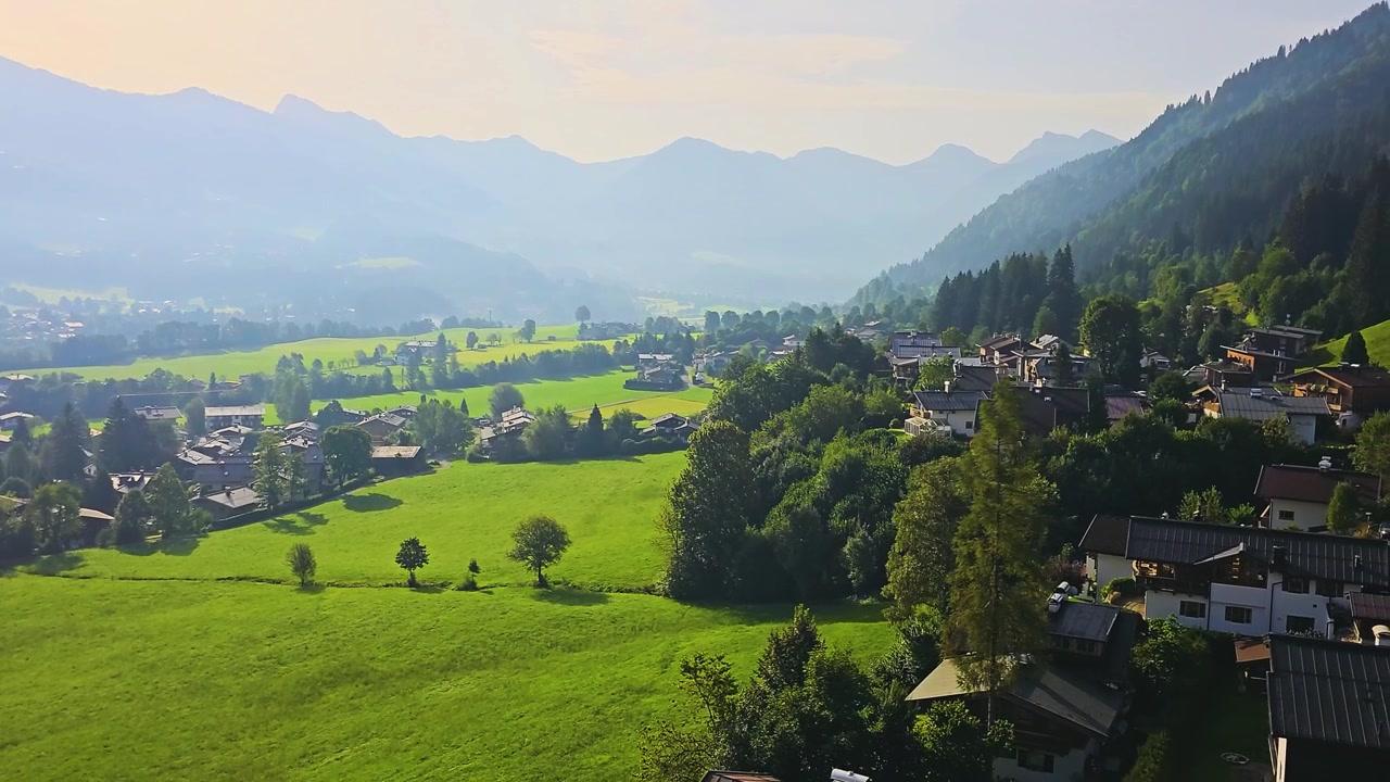 Vista dalla gondola in salita a Kitzbühel, Austria, che passa sopra tetti di villaggi, alberi, pascoli e pendii montani, con luce solare e foschia che creano una scena e un paesaggio di viaggio da sogno nel Tirolo