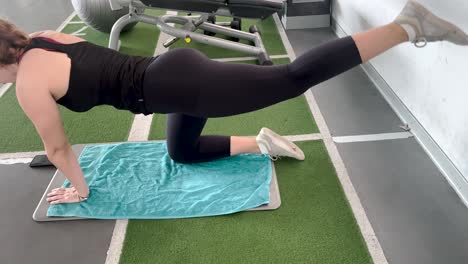 close view of young woman kneels to perform an exercise to work her back in a gym