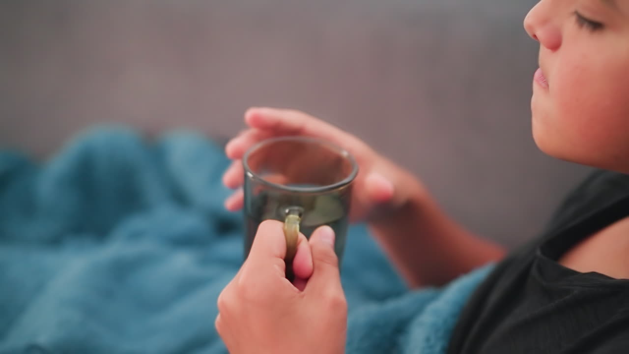 primer plano de un niño blanco sentado debajo de una manta tomando medicamentos con agua, centrado en la recuperación, el apoyo a la salud, la rutina de autocuidado y el bienestar infantil