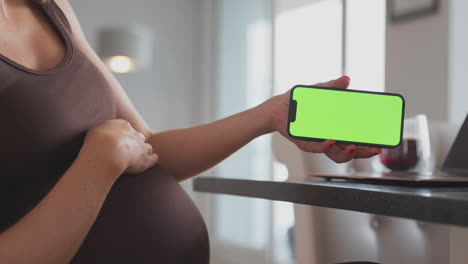 Nahaufnahme-Einer-Schwangeren-Frau,-Die-Zu-Hause-Am-Laptop-Arbeitet-Und-Ein-Mobiltelefon-Mit-Grünem-Bildschirm-In-Der-Hand-Hält
