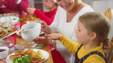 grandmother pouring gravy onto granddaughters food as multi-generation family eat christmas meal