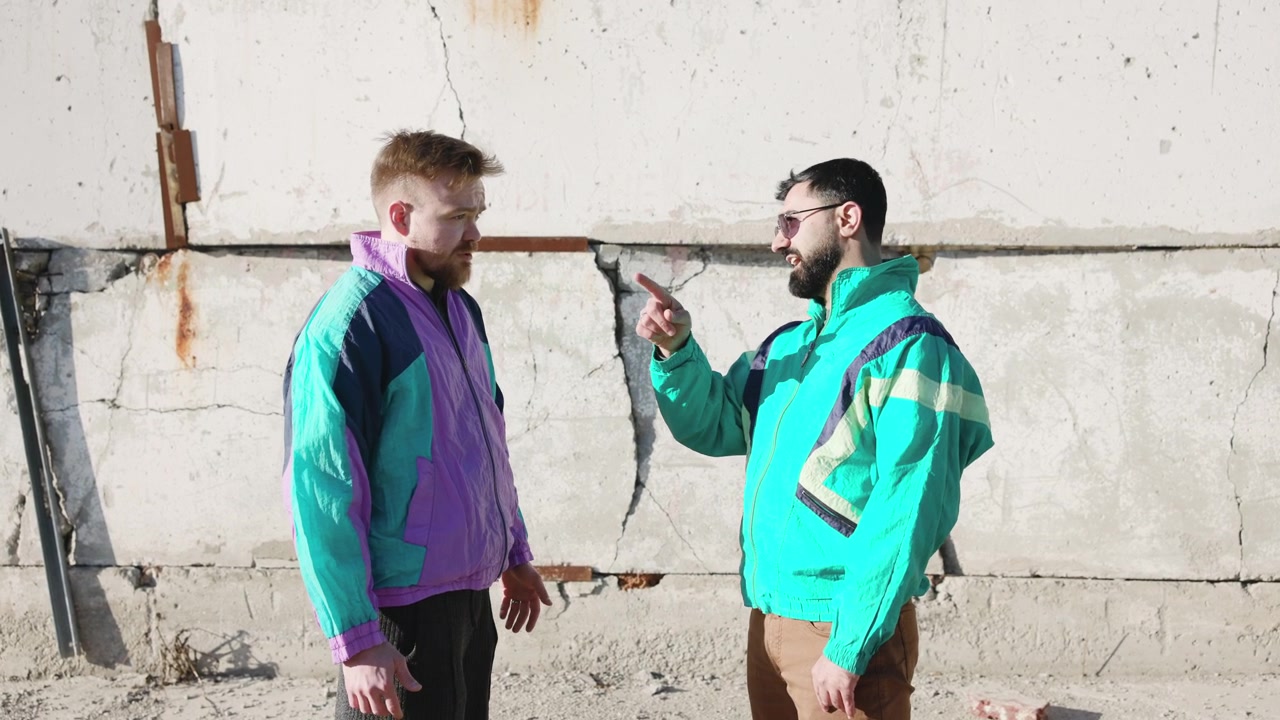 Two Men Confronting Near Concrete Wall, One Pointing Finger While Other Listens, Colorful Retro Windbreakers,