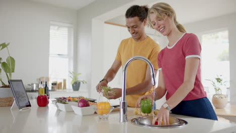 happy diverse couple preparing and washing fresh vegetables in kitchen with tablet, slow motion