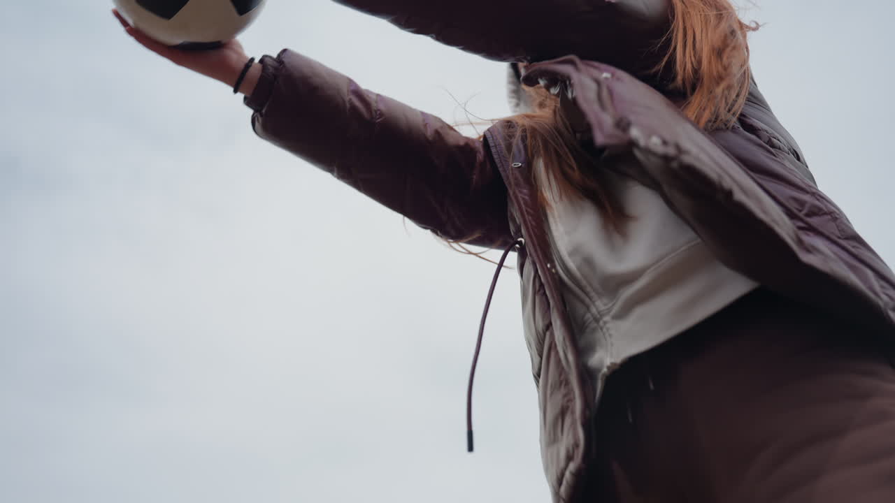 Primer plano de adolescente caucásico estirándose para alcanzar un balón de fútbol con guantes y chaqueta acolchada, foto de acción centrada que muestra las palmas, el pelo largo y gorro, cielo de fondo y luz invernal nítida en campo urbano