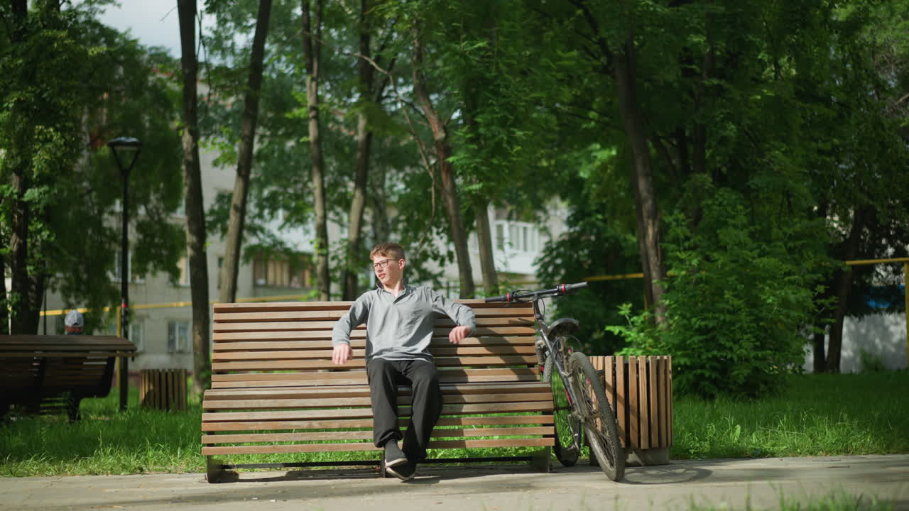 White boy looks back before sitting on wooden bench outdoors, he stretches, placing his right foot over his left, a bicycle is parked next to him, with another person seated on a distant bench