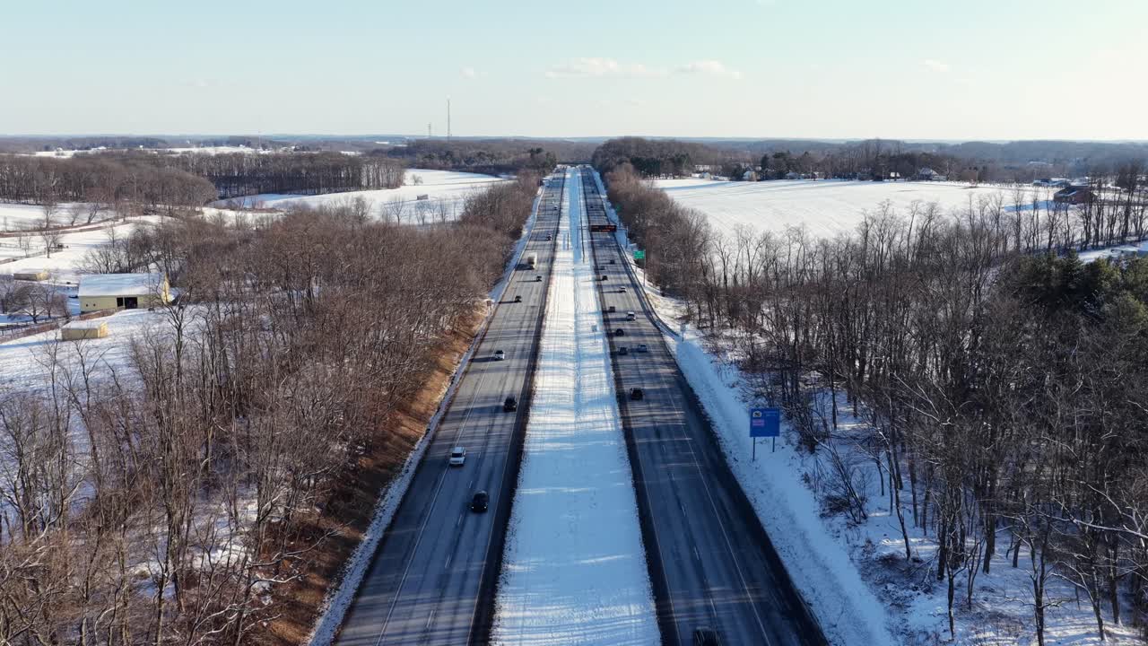 Vista aerea di una lunga strada statale rurale che attraversa terreni agricoli innevati in America, con traffico leggero che scorre tra campi ghiacciati e alberi invernali spogli sotto la luce chiara del giorno. Inquadratura ampia. Confine PA MD.