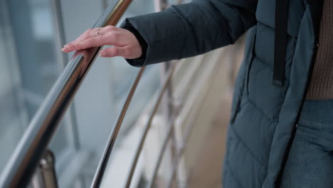 close-up of lady hand in black hoodie with ring-adorned fingers gliding on sleek metal rail as she walks, with reflective building glass showing outdoor cityscape in soft, natural light