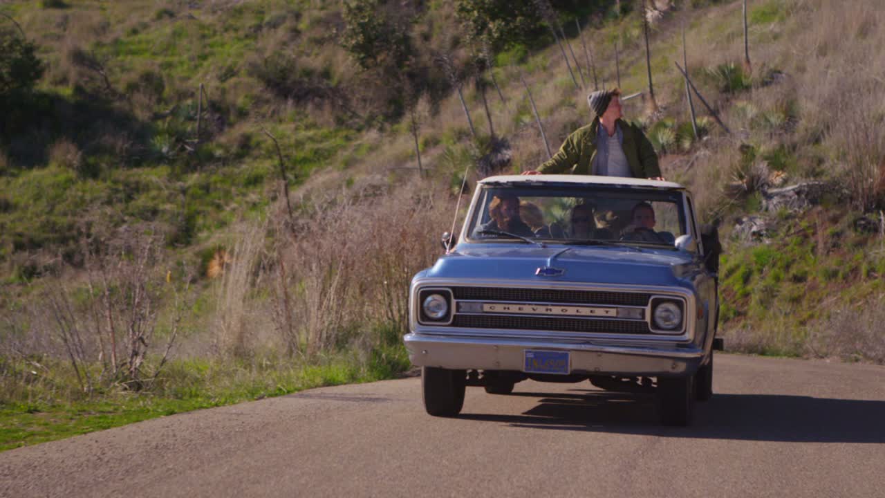 un grupo de amigos en una camioneta azul conducen por una carretera rural mientras un hombre en la camioneta se para
