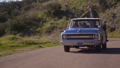 a group of friends in a blue pickup truck drive on a rural road while a man in the truck bed stands