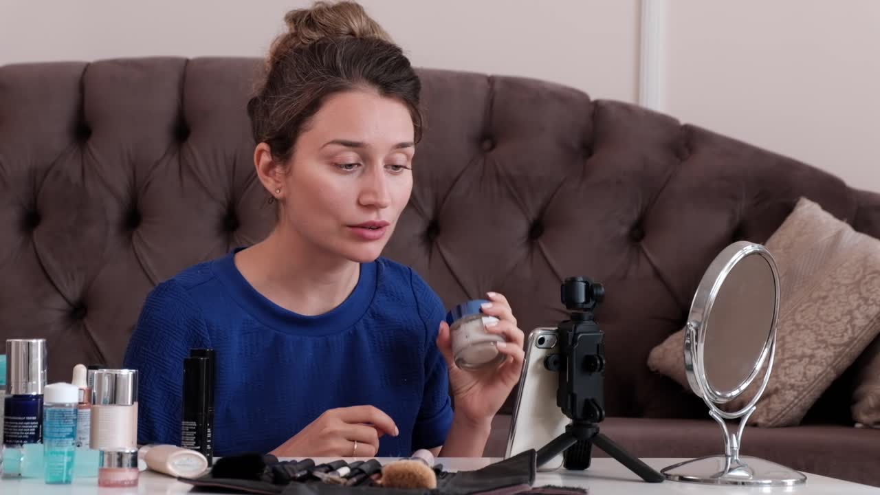 Woman in a blue T-shirt filming herself while doing a make-up tutorial at home