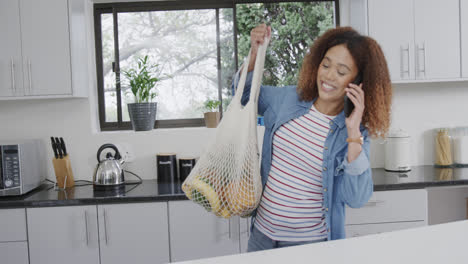 Feliz-Mujer-Birracial-Hablando-Por-Teléfono-Dejando-Bolsas-De-Compras-En-La-Cocina,-Cámara-Lenta